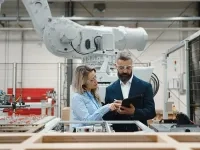 Deux personnes discutent et consultent une tablette dans une usine automatisée, devant un grand bras robotisé.